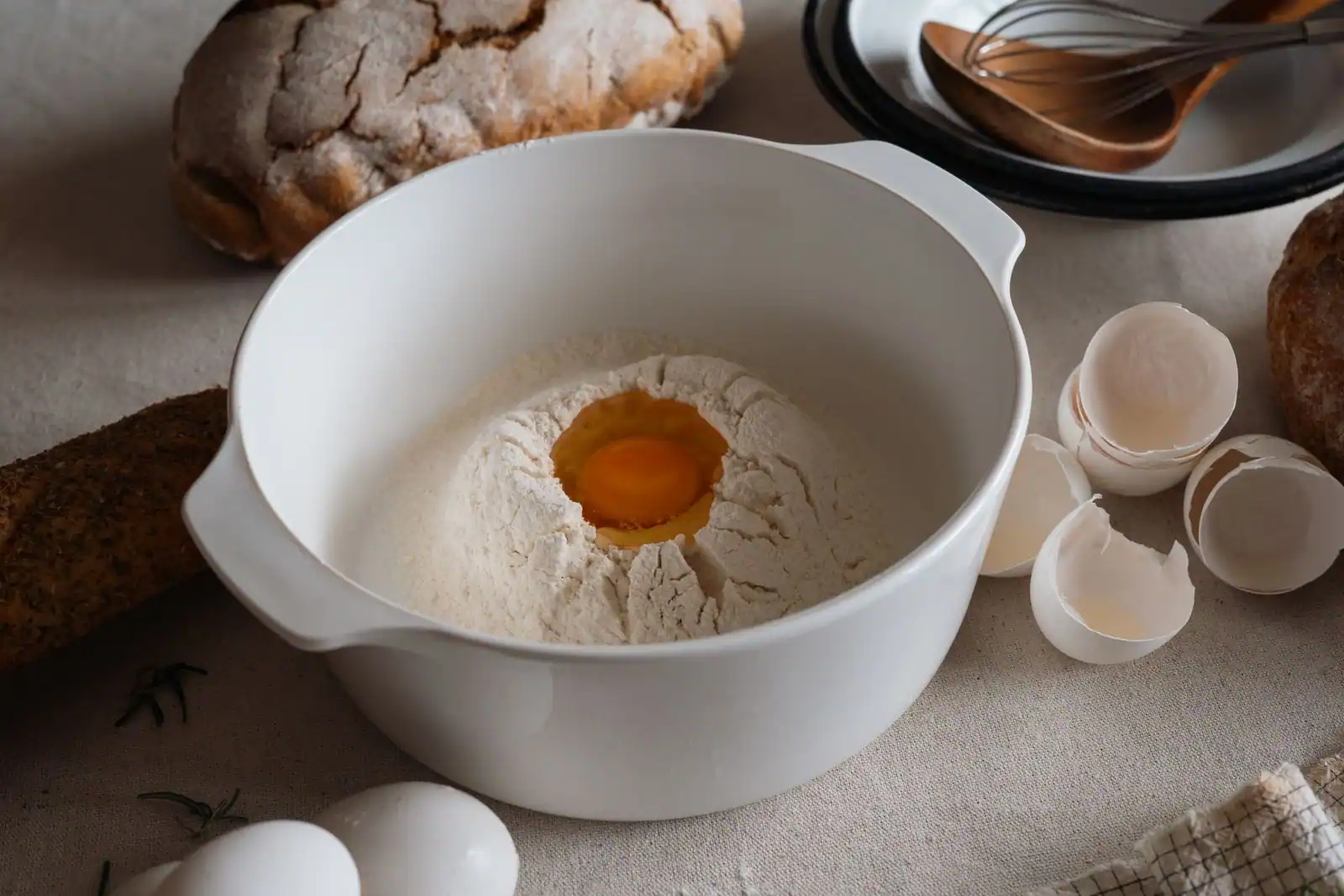Bol blanc rempli de farine avec un jaune d'œuf et un fouet, entouré de pains et d'œufs sur une table