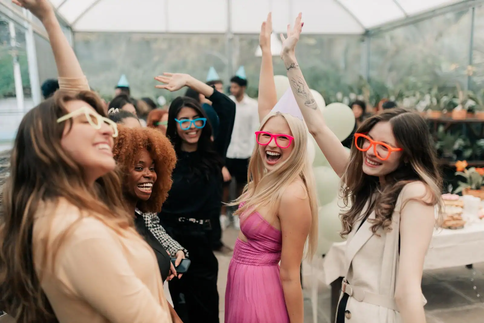 Des amies adultes rient et lèvent les bras lors d'une fête, avec chapeaux et lunettes colorés.
