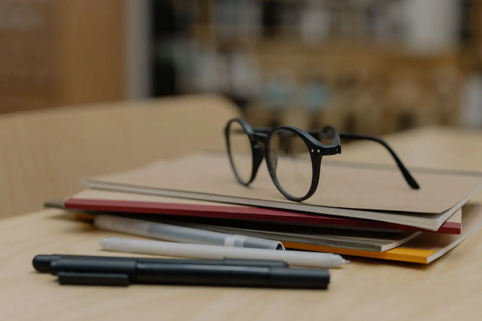 Lunettes rondes posées sur une pile de carnets, avec des stylos, sur une table en bois claire.