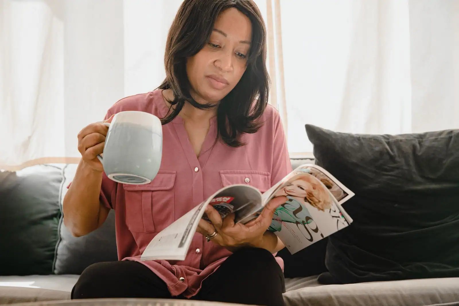 Une femme lit un magazine lifestyle chez elle, une tasse de café à la main.