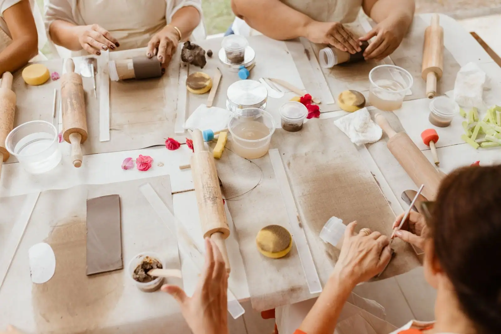 Vue de dessus d'un atelier de loisirs créatifs en groupe : plusieurs mains travaillent l'argile et des matériaux autour d'une table.