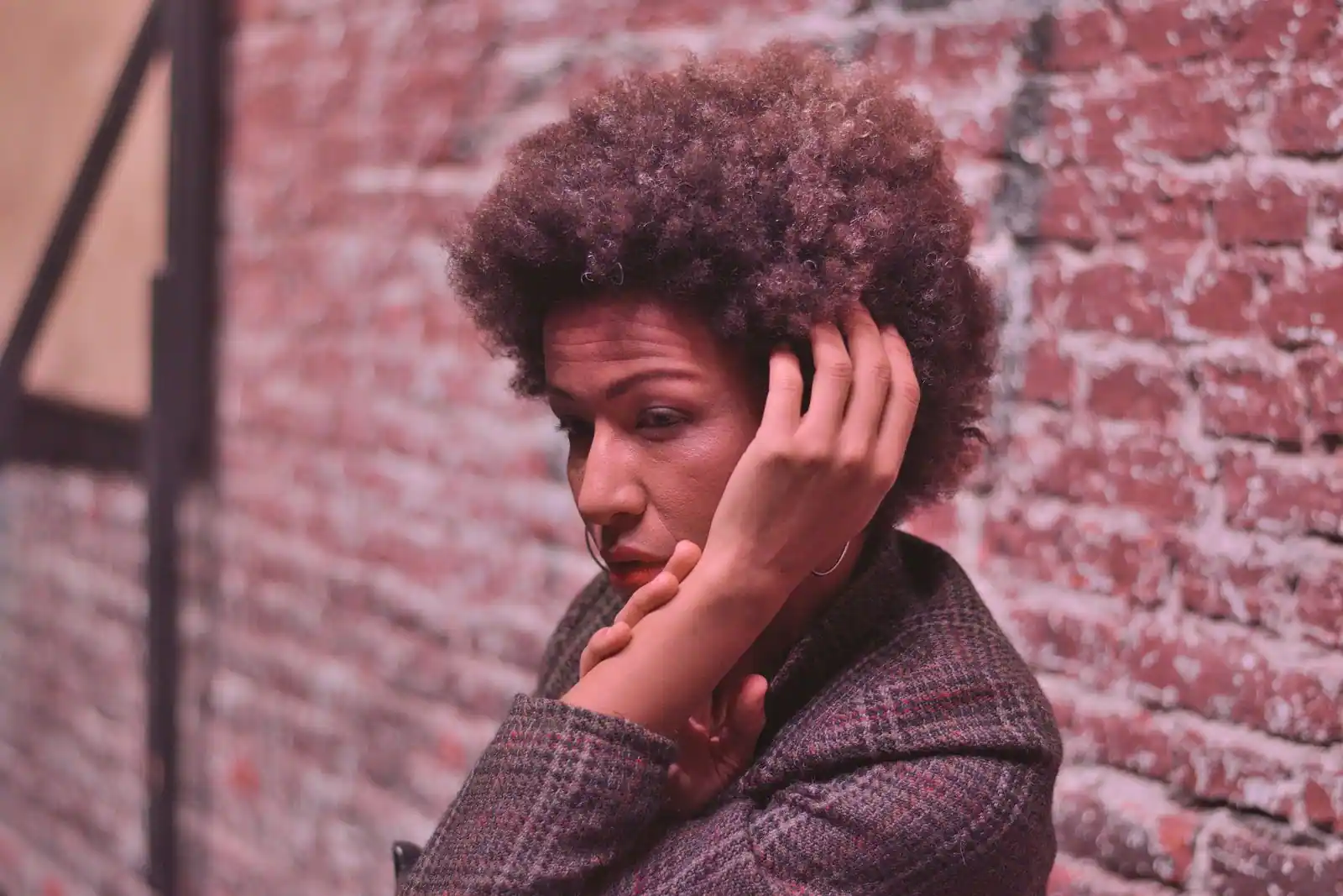 Femme aux cheveux crépus afro, une main dans les cheveux, devant un mur de brique