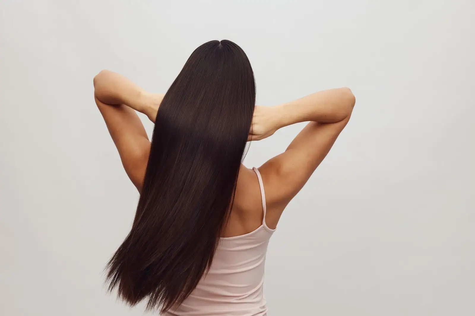 Une femme vue de dos dévoile de longs cheveux lisses et soignés sur un fond clair uni