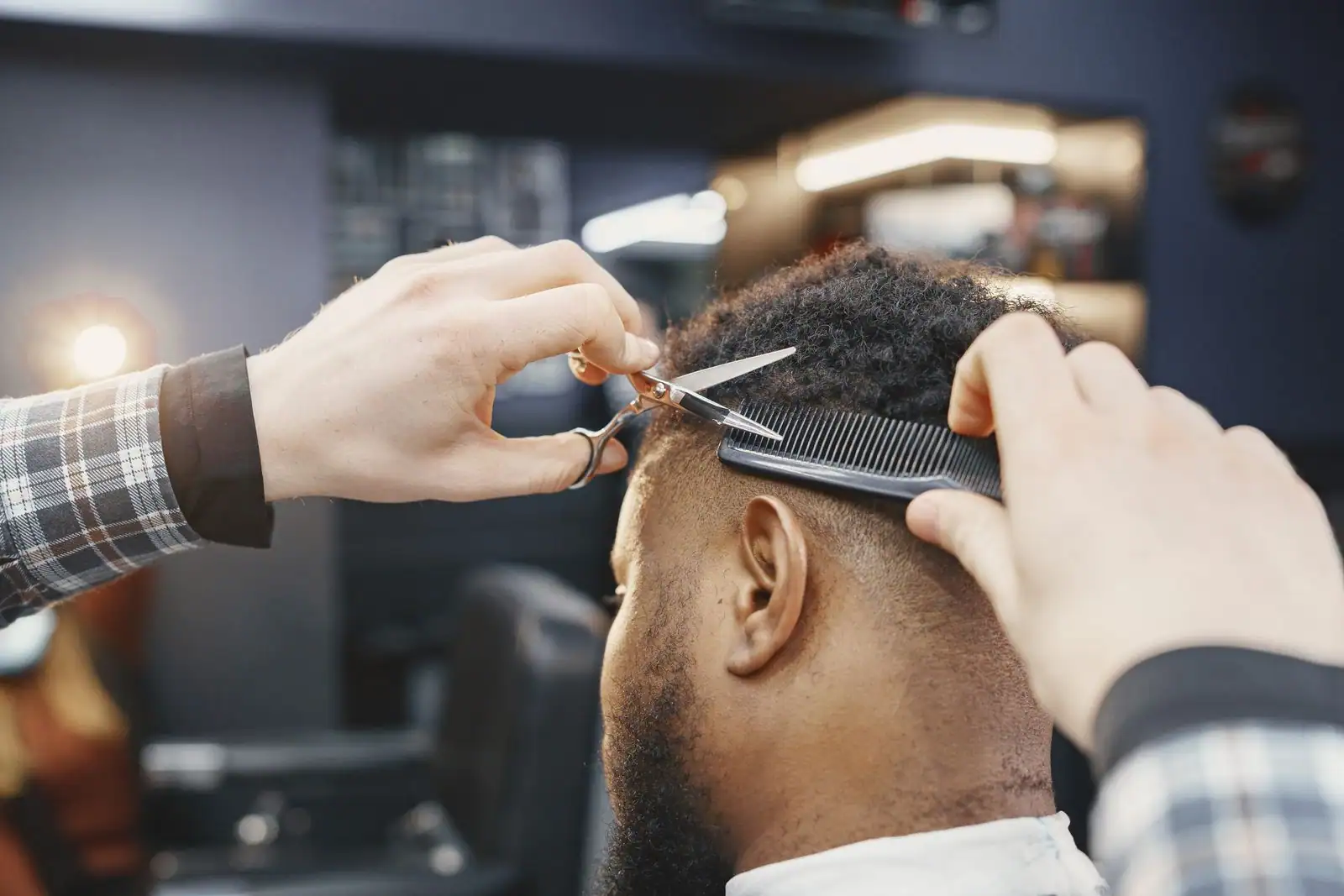 Un barbier coupe aux ciseaux les cheveux d'un homme vu de dos, peigne en main, en salon