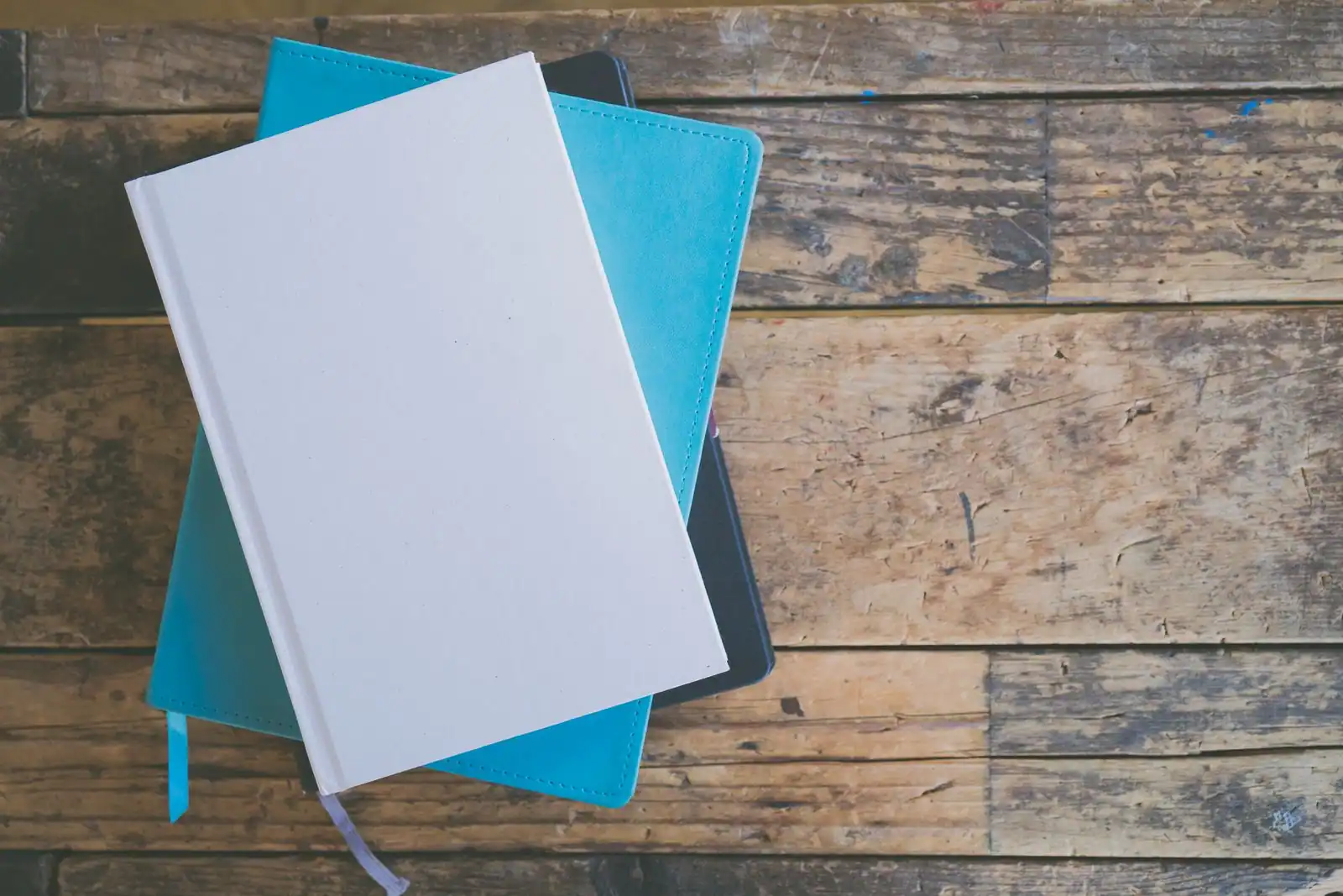 Pile de carnets fermés, un blanc sur un turquoise, posés sur une table en bois rustique vue de dessus.