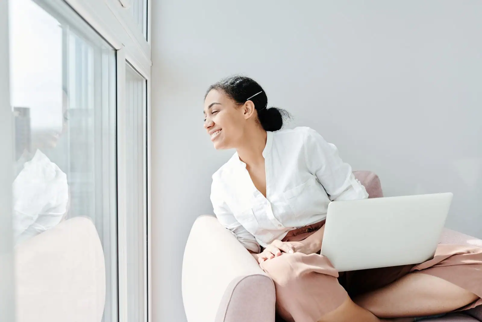 Femme souriante assise près d'une fenêtre avec un ordinateur portable sur les genoux