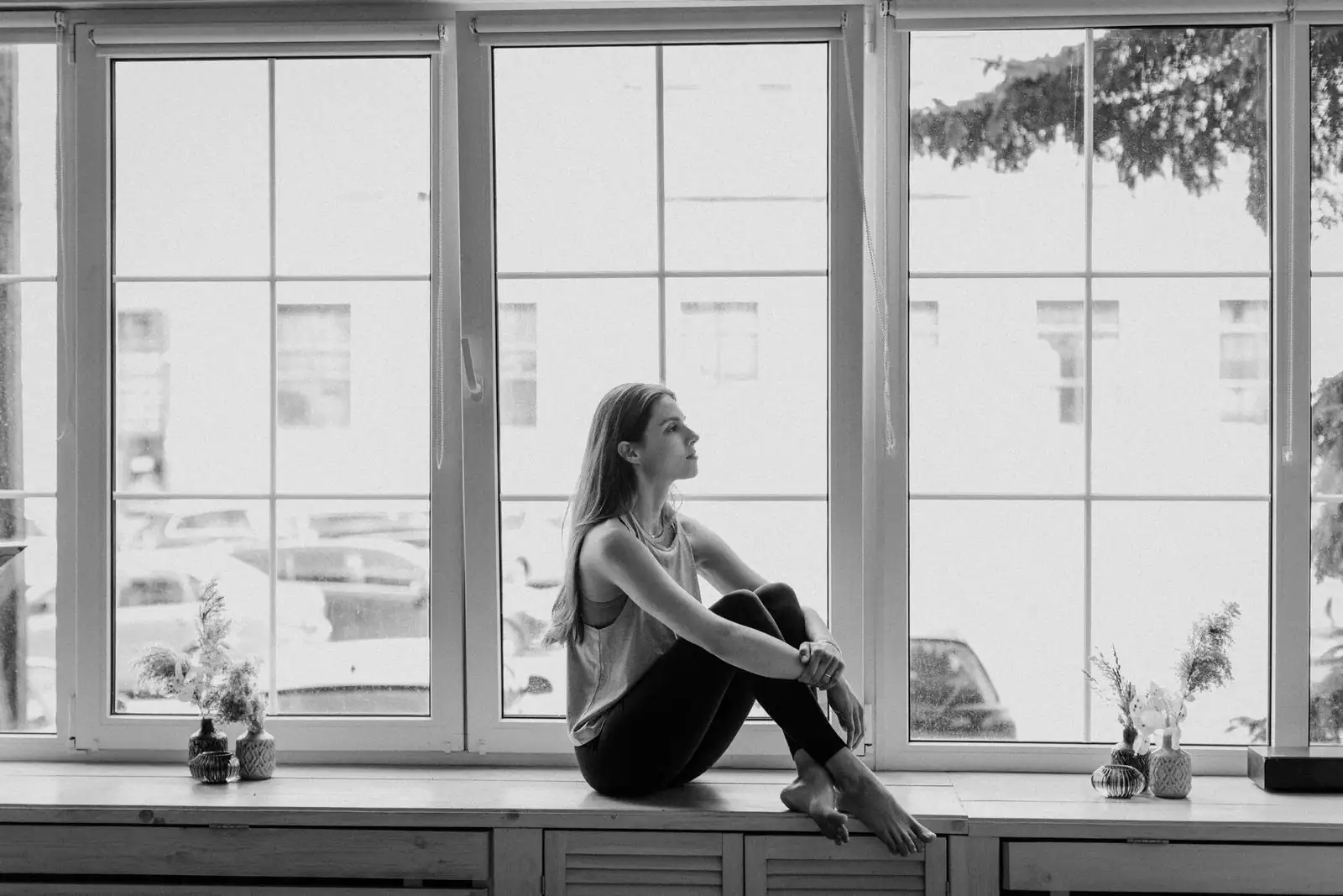 Photo en noir et blanc d'une jeune femme pensive assise sur le rebord d'une grande fenêtre, regardant dehors.