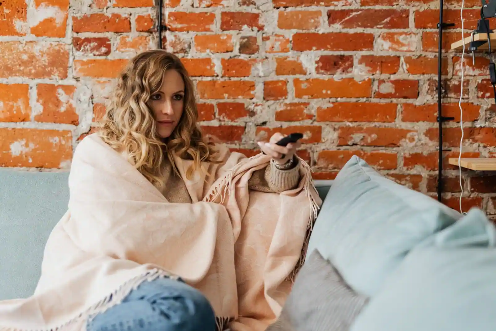 Femme enveloppée dans un plaid sur un canapé, tenant une télécommande de télévision