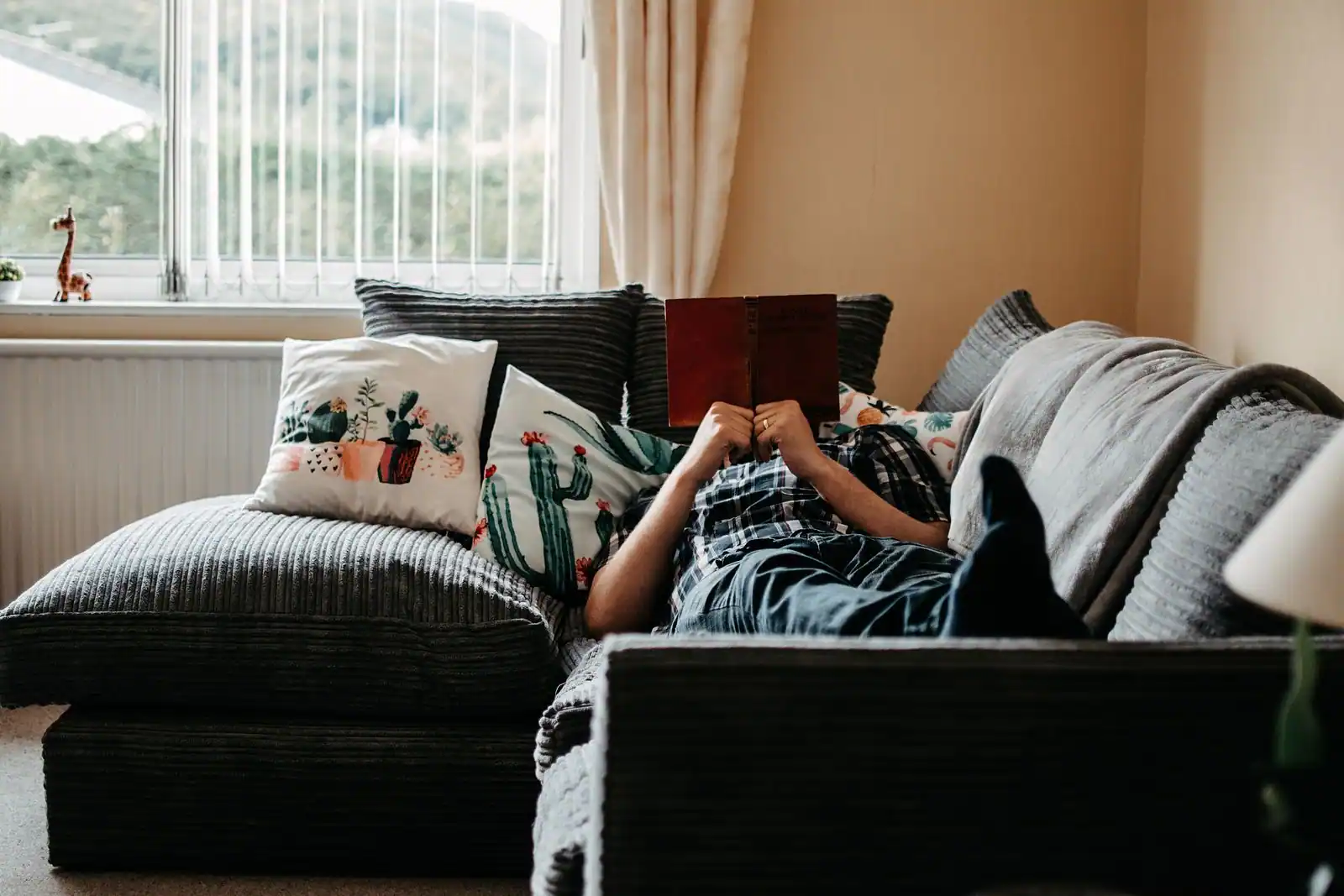 Personne allongée sur un canapé moelleux, lisant un livre près d'une fenêtre, dans une ambiance calme et cosy du quotidien.