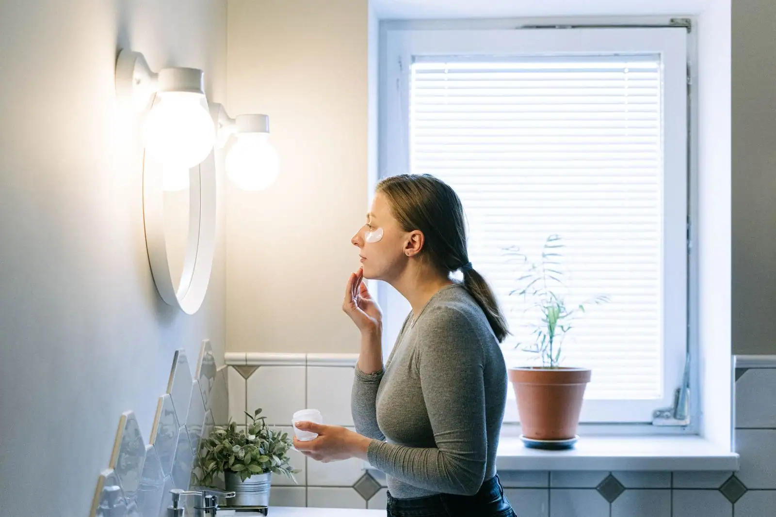 Femme en tee-shirt gris appliquant une crème du bout des doigts sur la joue, un pot à la main, dans une salle de bain.
