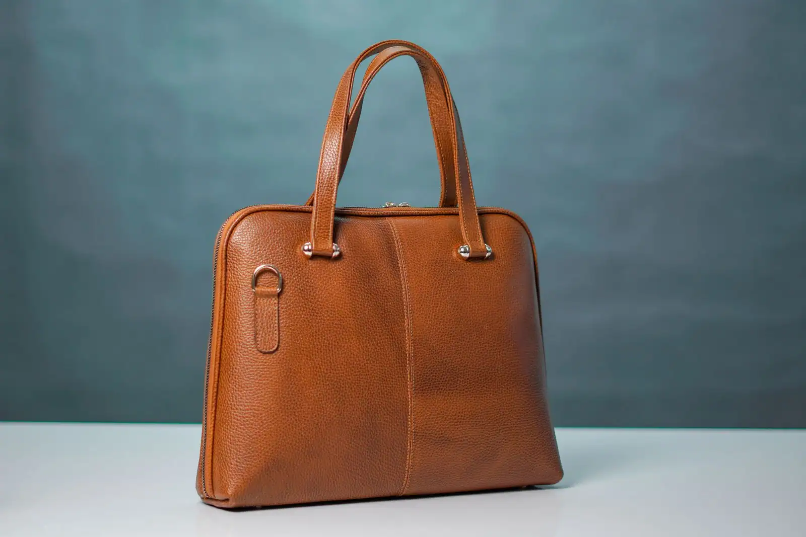 Sac à main en cuir grainé cognac à deux anses, posé sur une surface claire devant un fond gris-vert épuré.