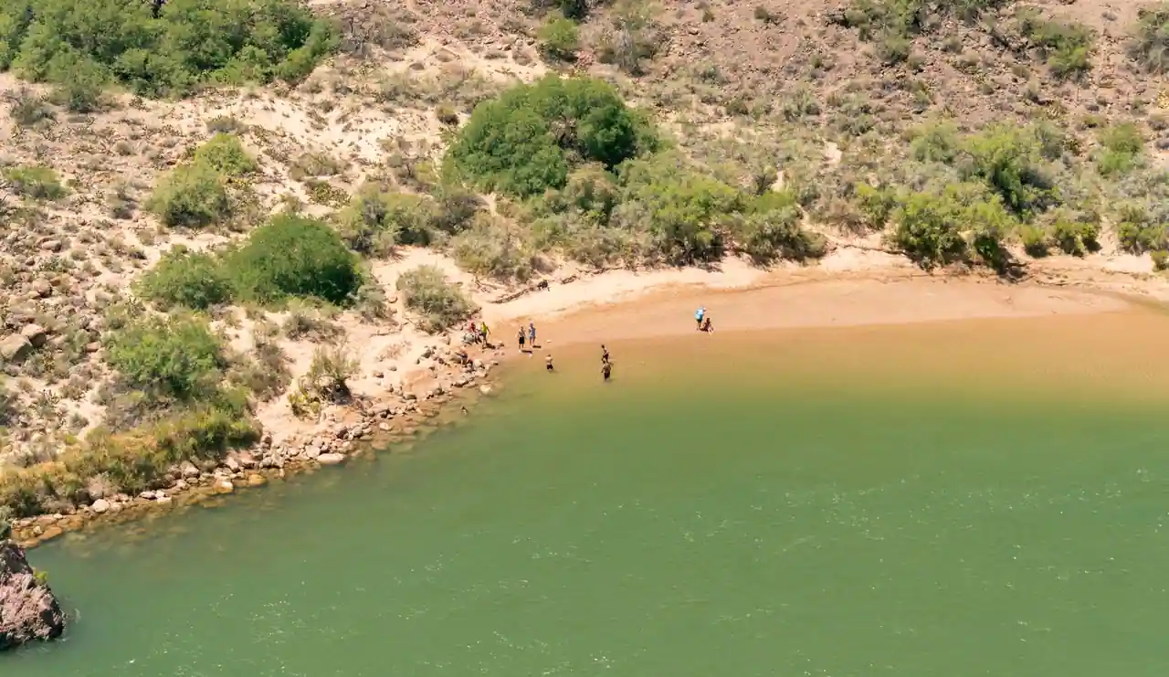 Plage de rivière aux eaux vertes bordée de collines arides, avec quelques personnes au bord de l'eau l'été