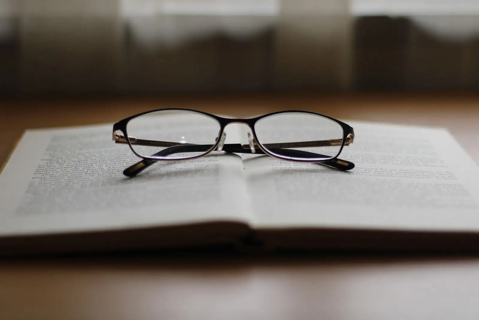 Livre ouvert et lunettes de lecture posés sur une table en bois, fenêtre floutée en arrière-plan, lumière naturelle douce