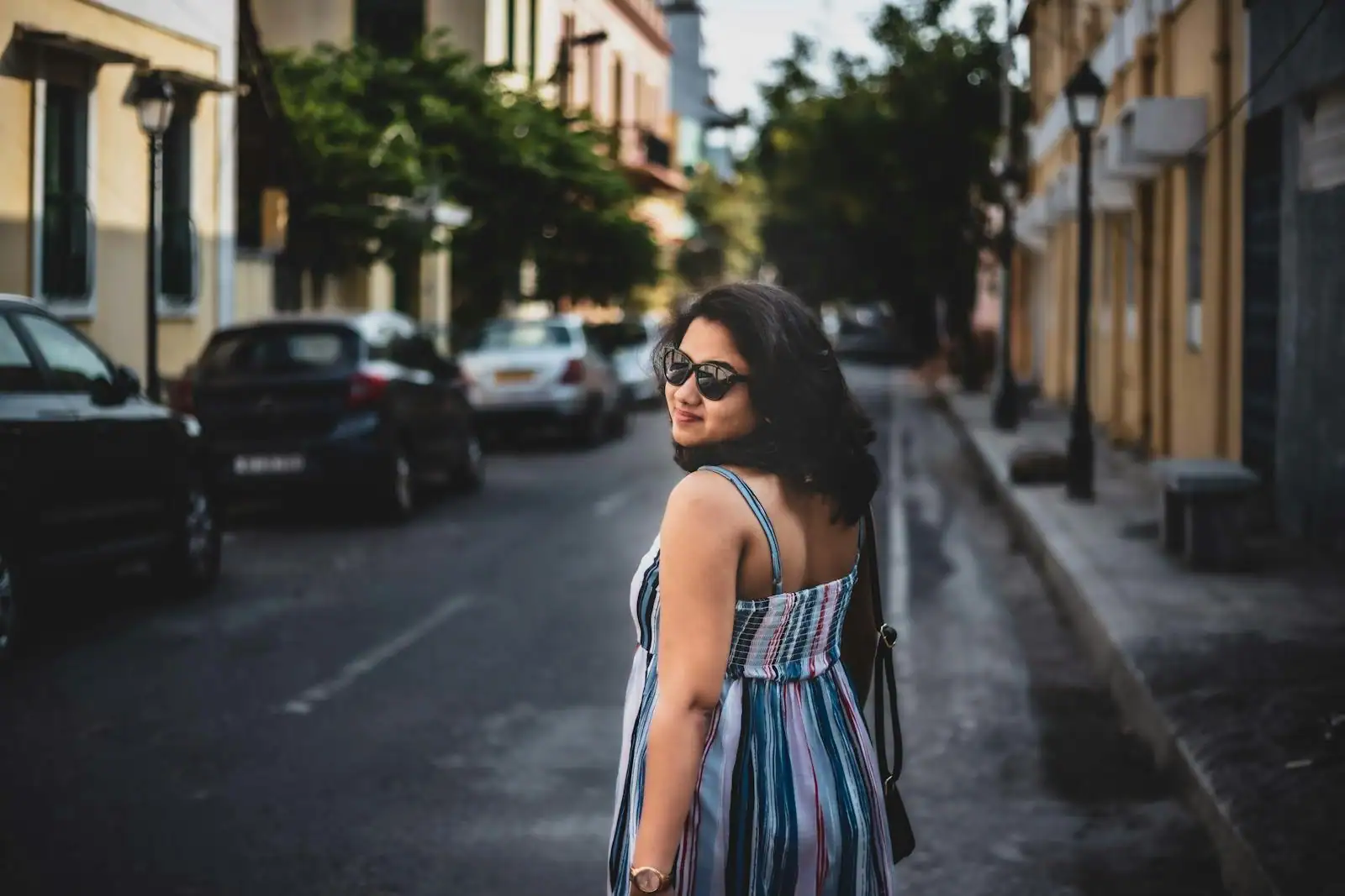 Femme en robe d'été rayée et fluide dans une rue ensoleillée, lunettes de soleil, allure de saison décontractée