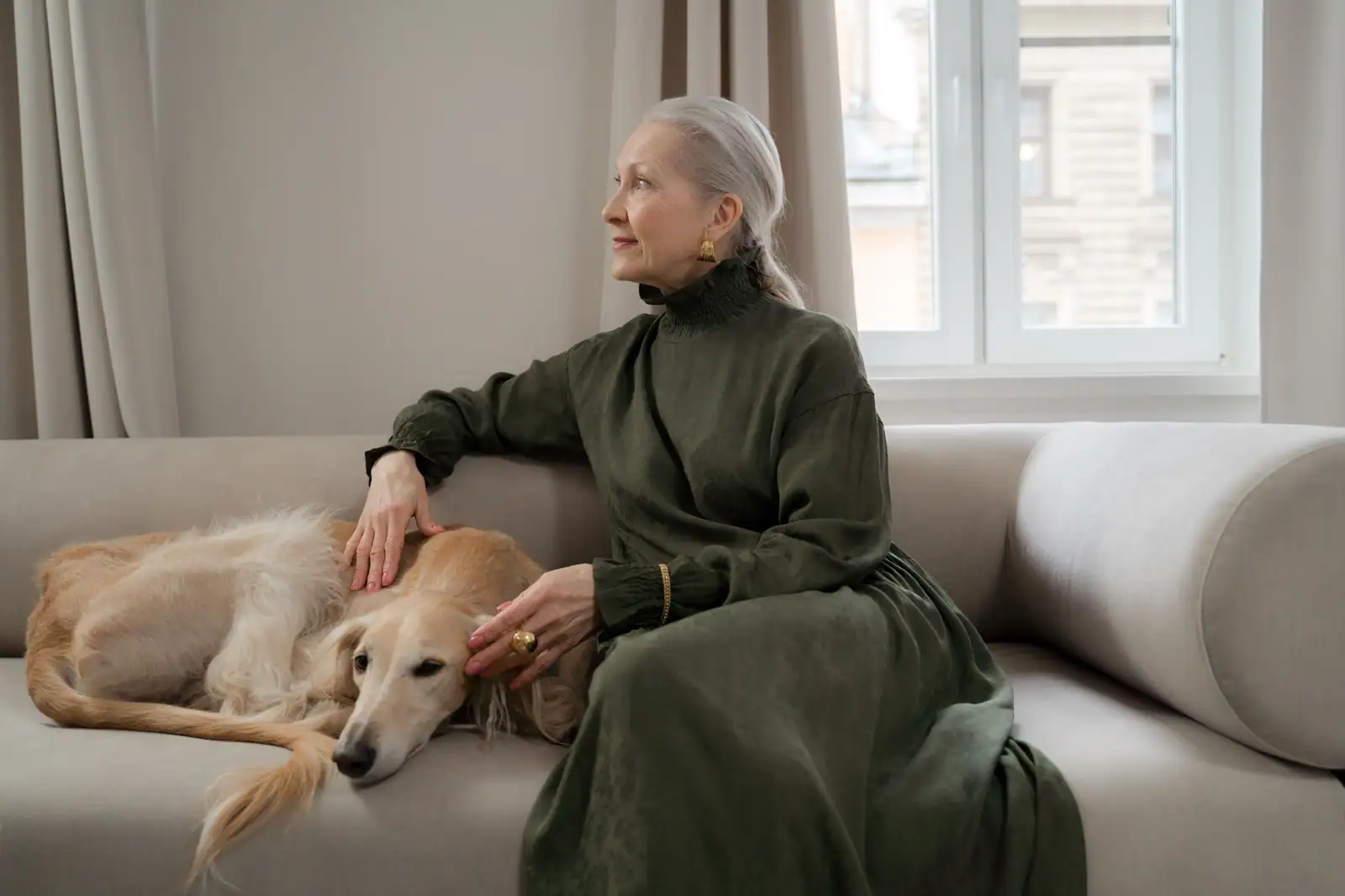 Femme installée sur un canapé, la main posée sur son lévrier couché contre elle près de la fenêtre.