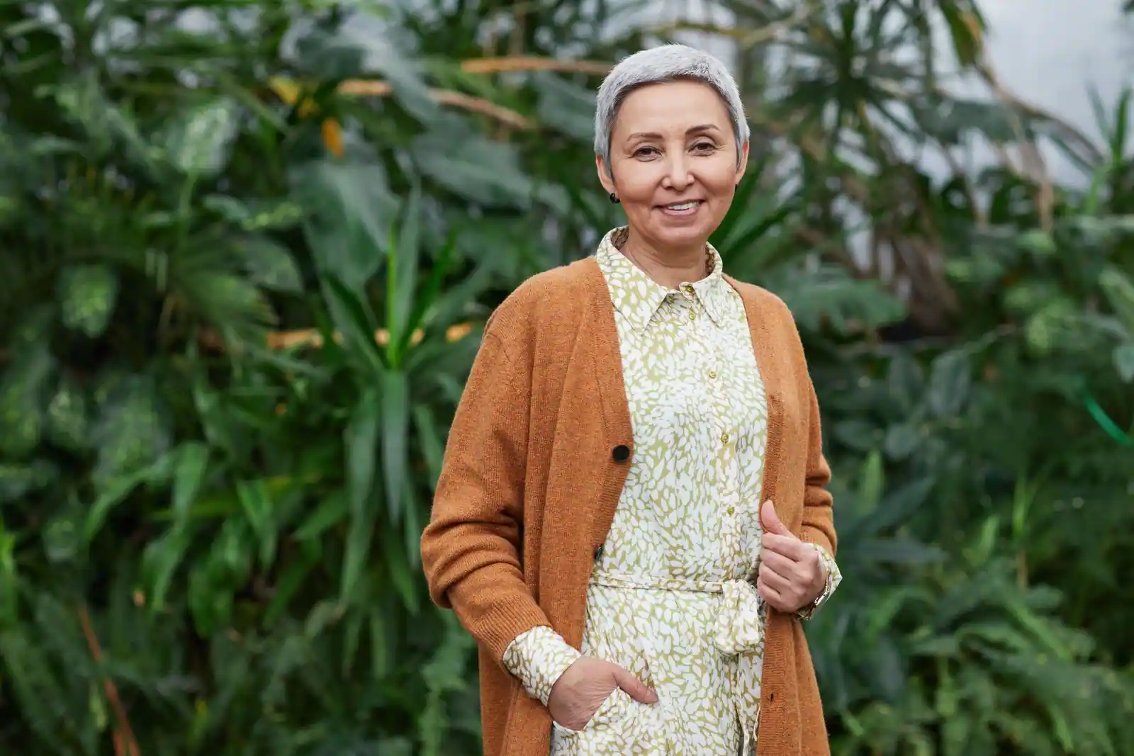 Femme aux cheveux gris argentés coupés court, souriante devant un feuillage vert en extérieur