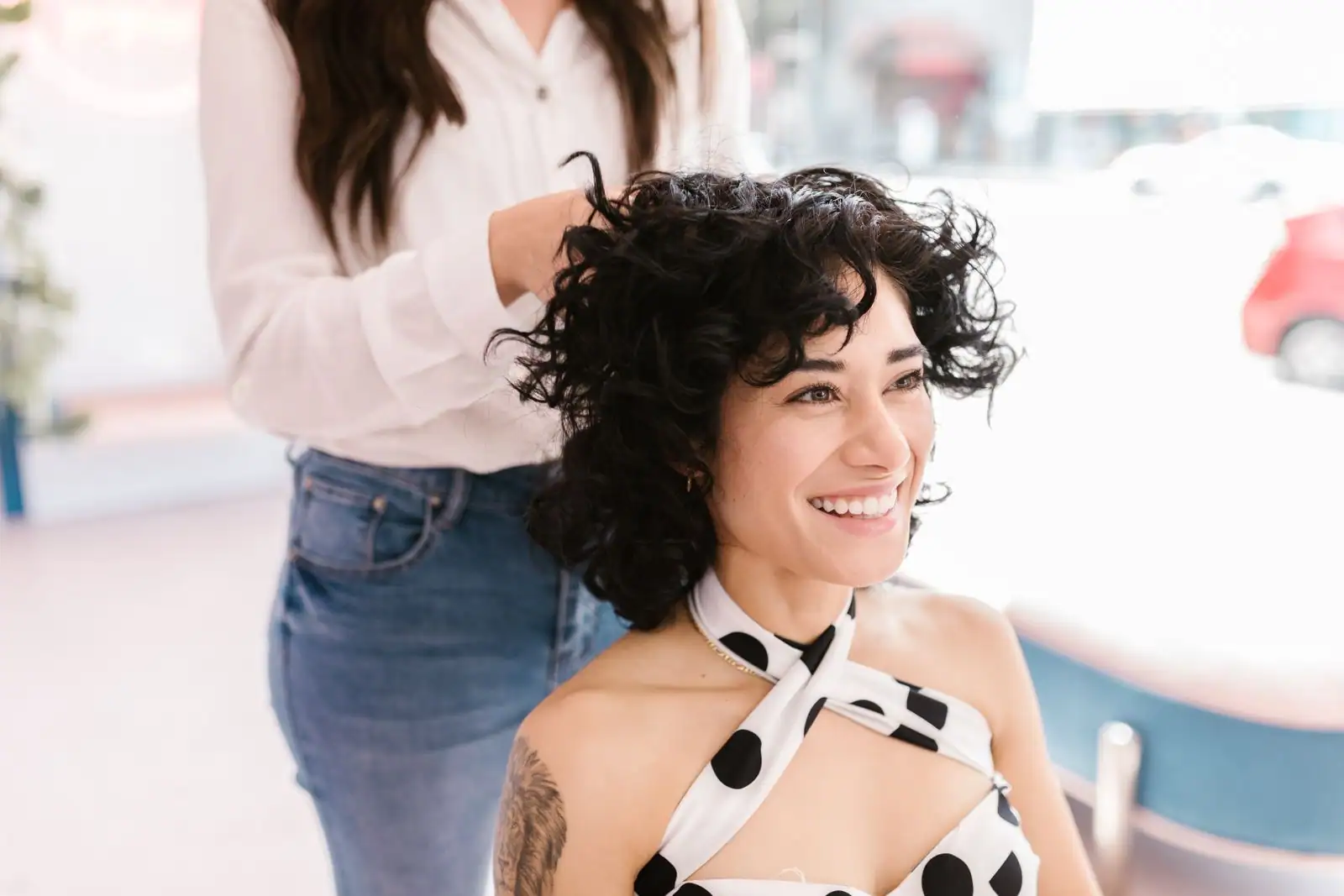 Femme aux cheveux courts très bouclés dans un salon, coiffée par une professionnelle derrière elle