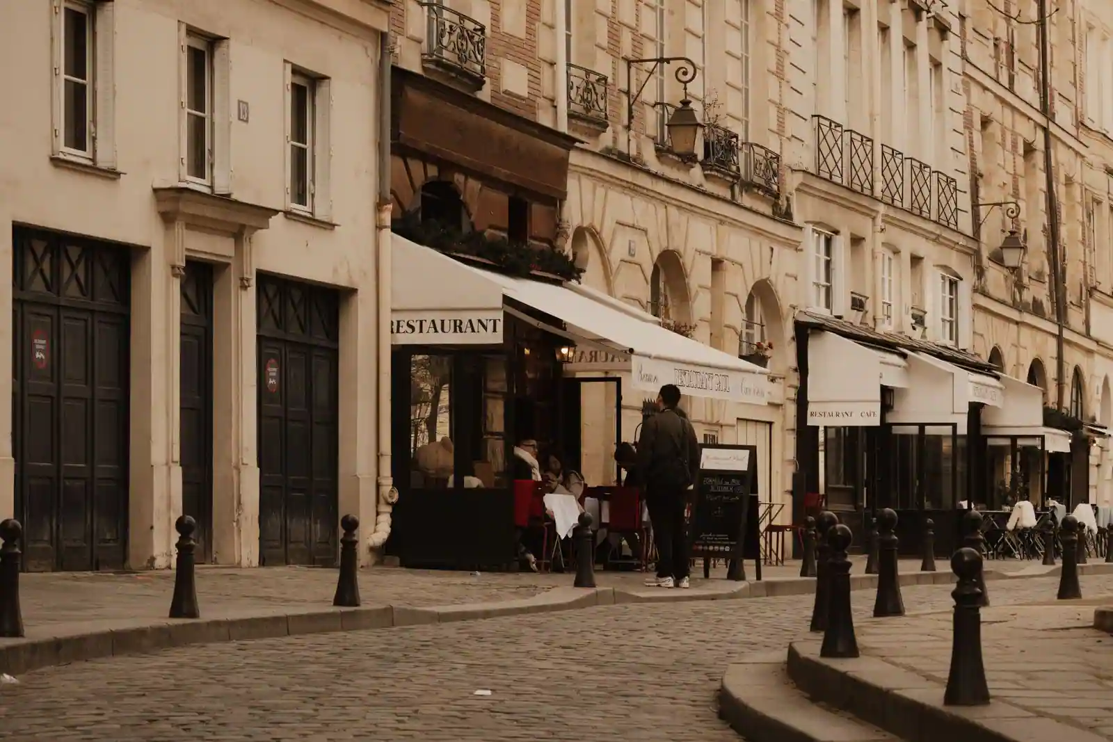 Rue pavée parisienne bordée de terrasses de restaurant, avec une personne devant une ardoise de menu