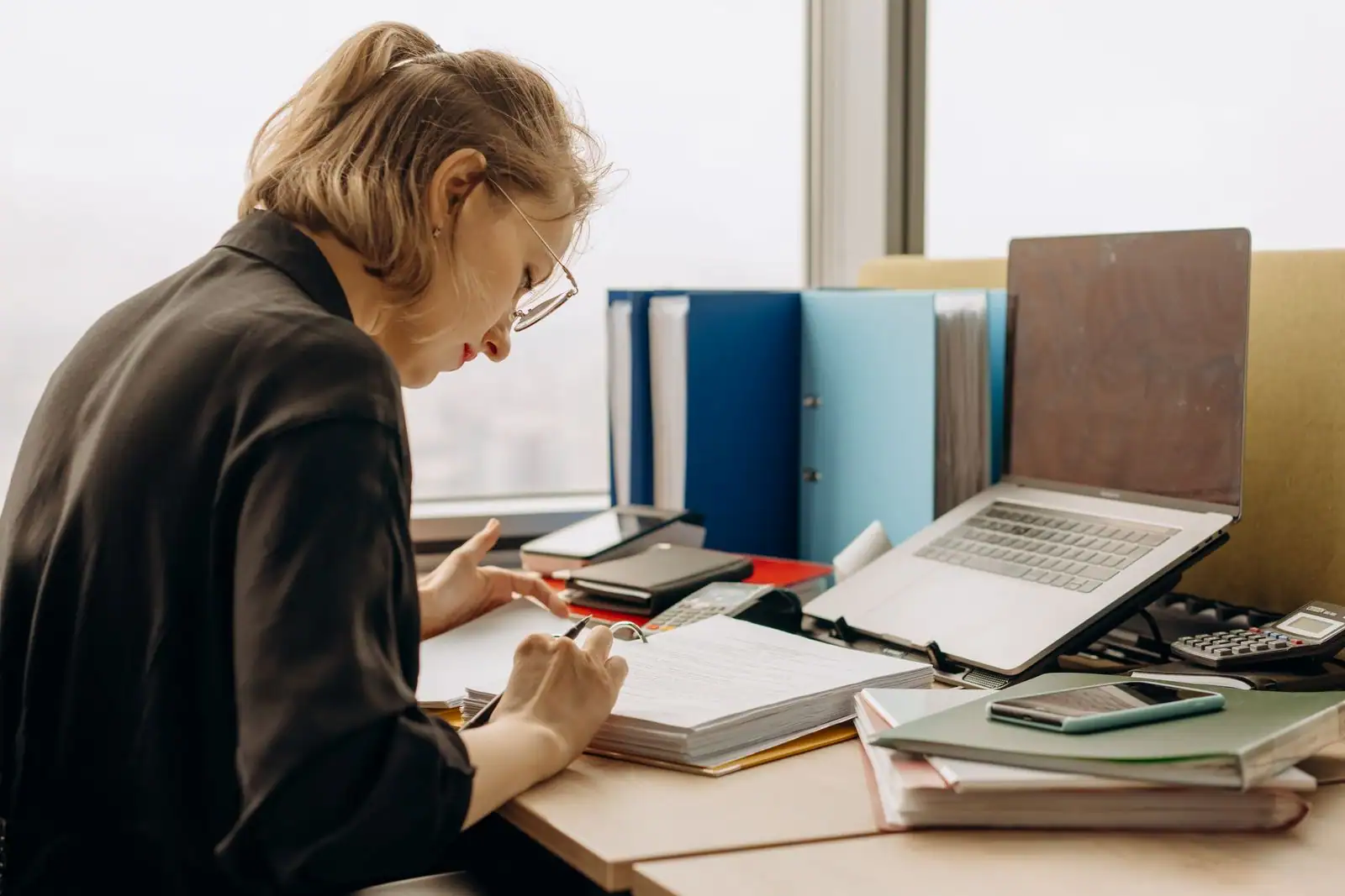 Une femme annote des documents dans un classeur, à un bureau couvert de dossiers et d'un ordinateur.