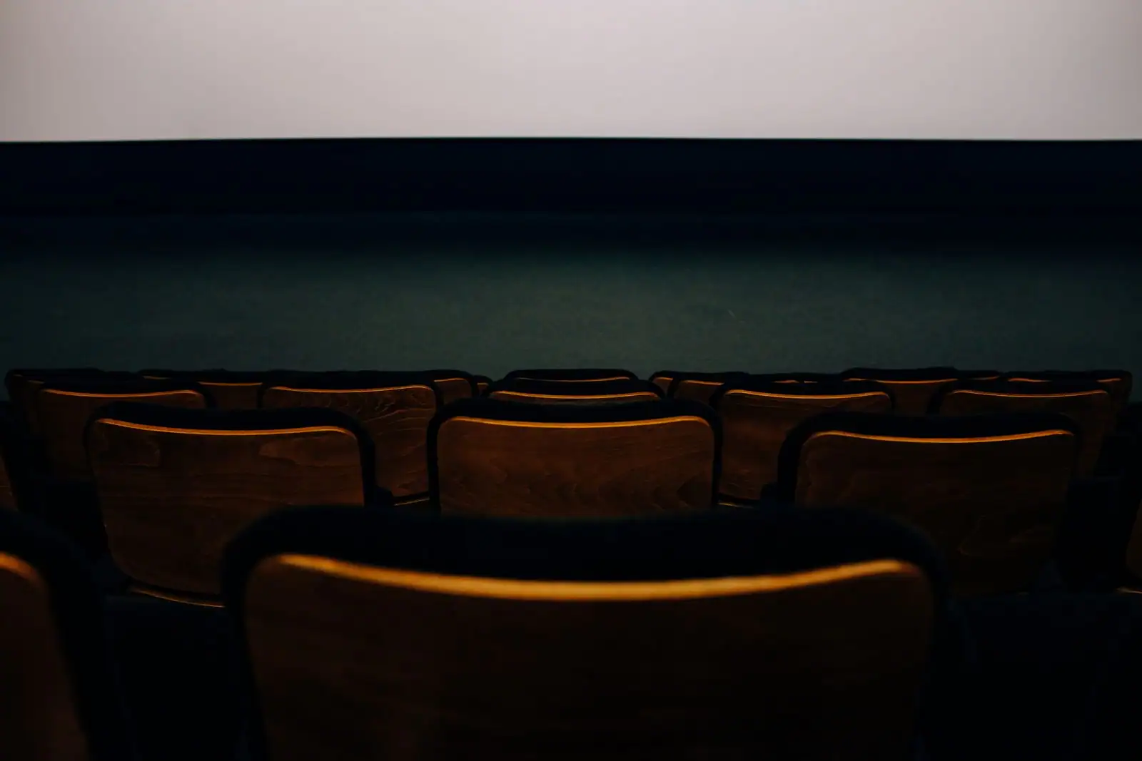 Salle de cinéma déserte, rangées de fauteuils en bois sombre tournées vers un écran éclairé.