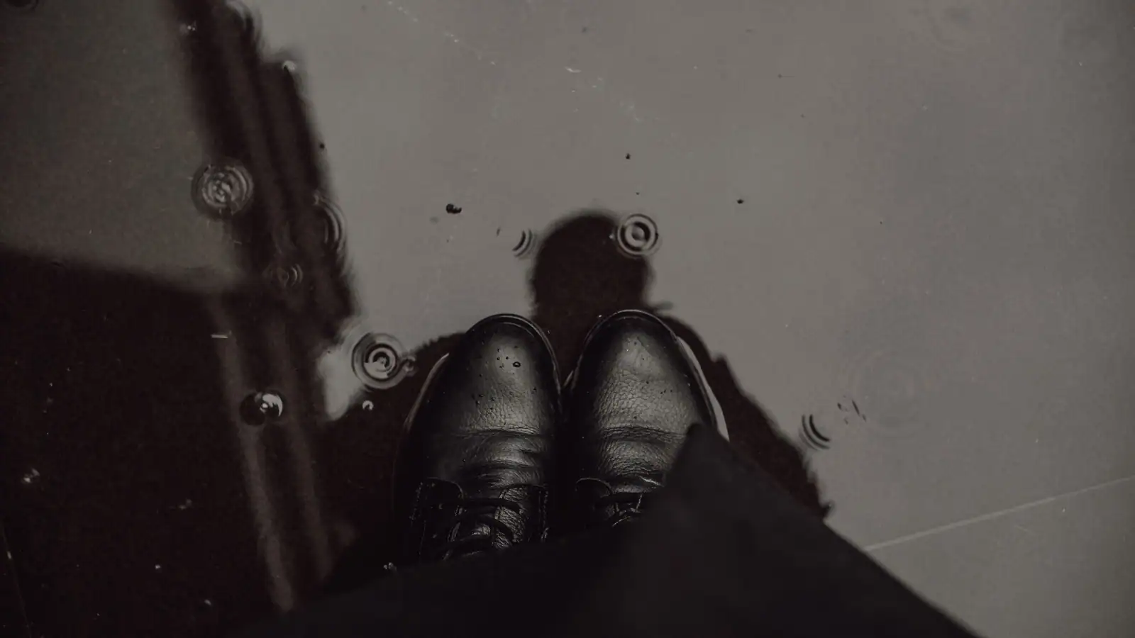 Chaussures en cuir noir et bas d'un vêtement sombre, vus de haut sur un sol mouillé de pluie