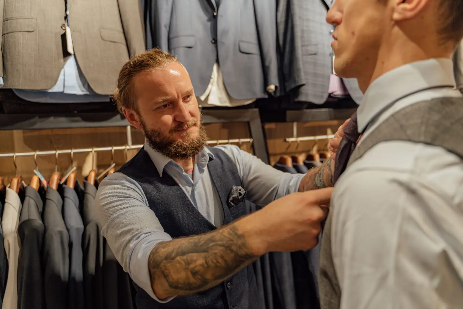 Homme à la barbe soignée et bien contourée, en gilet et chemise, ajustant le col d'une tenue chez un tailleur