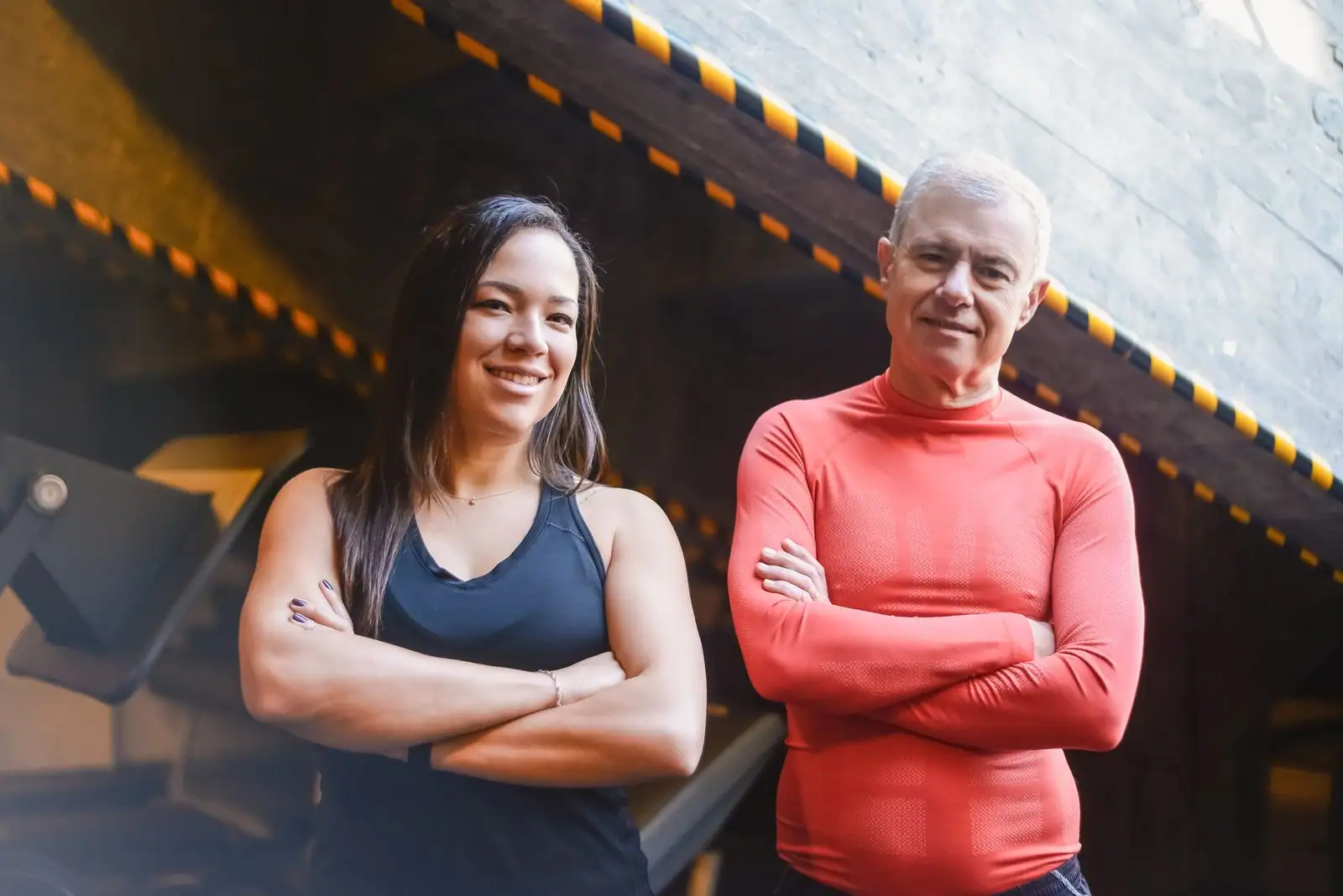 Deux personnes debout, bras croisés, souriantes et à l'attitude confiante dans une salle de sport.