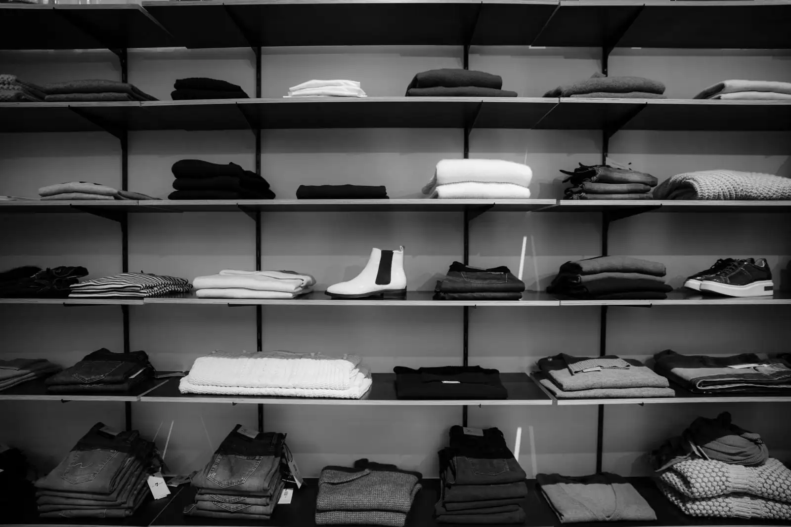 Étagères de vêtements pliés et chaussures présentées en boutique, photo en noir et blanc.